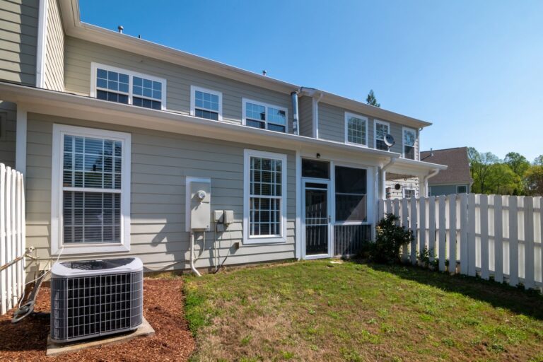 Outdoor air conditioning condenser unit on the lawn beside a suburban home with white fencing
