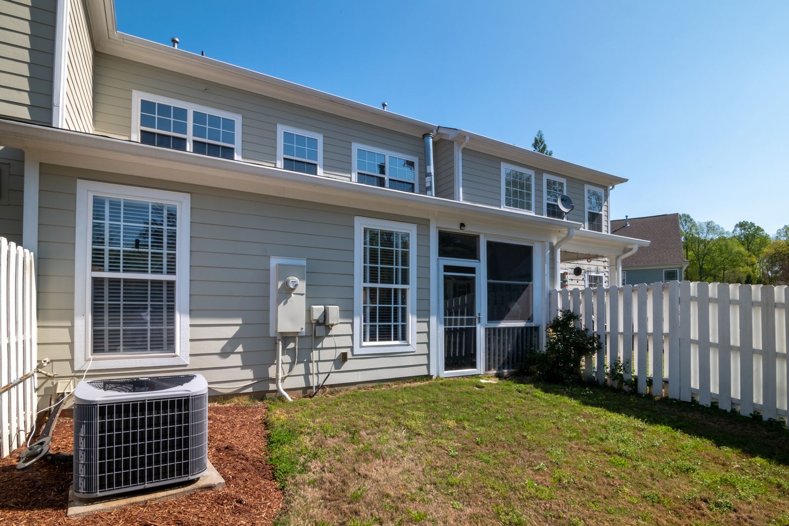 Outdoor air conditioning condenser unit on the lawn beside a suburban home with white fencing